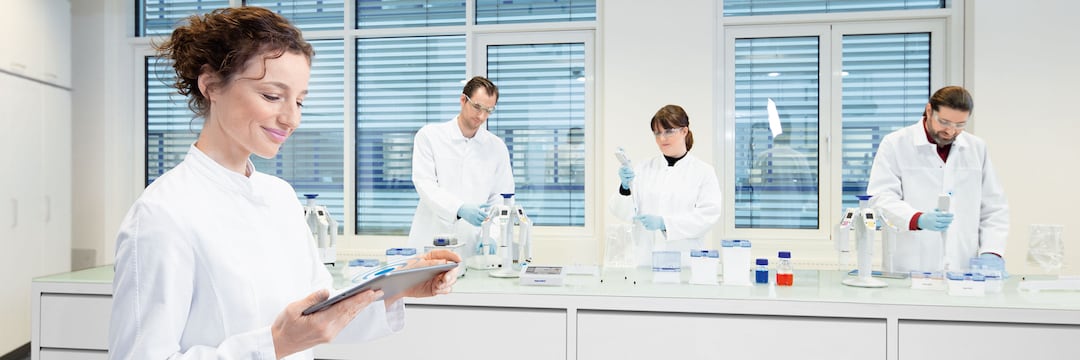 Female lab assistant holding tablet against the background of a laboratory setting with 3 colleagues working with connected pipettes.