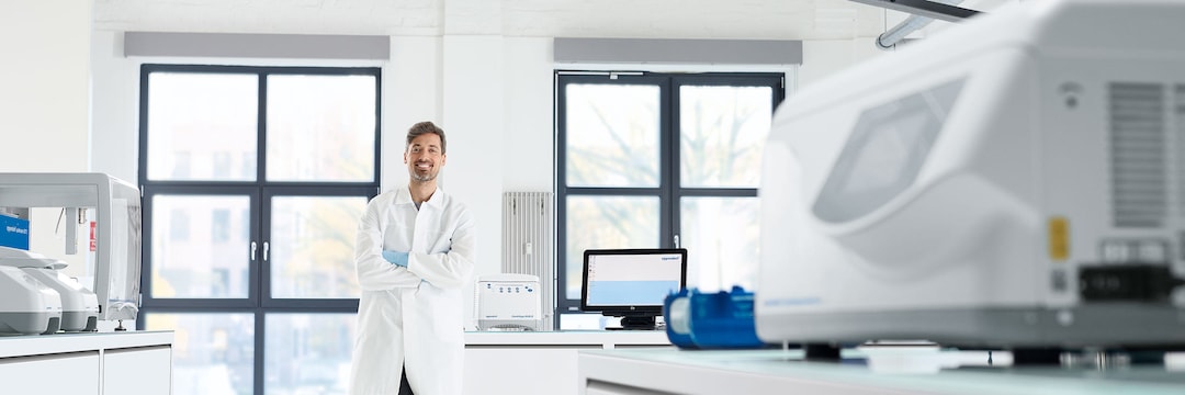 Male laboratory assistant standing in a laboratory room with Centrifuges at the back of a window, arms folded in front of chest and smiling contentedly.