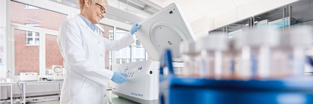 Lab scene with female laboratory assistant in the background who closes the lid of an Eppendorf Centrifuge 5920 R and another female laboratory assistant sitting behin a lab desk and dispensing red liquid in a plate